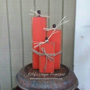 May include: Three orange wooden pumpkins stacked on top of each other. The pumpkins are tied together with twine and have a rustic look. The pumpkins are sitting on top of a rusty metal container. The text "Kelly's Unique Primitives" and "www.kellyuniqueprimitives.com" is visible on the container.