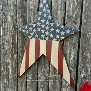 Op de afbeelding: Een houten stervormig bord geschilderd met de Amerikaanse vlag. De ster is geschilderd met rode en witte strepen en blauwe sterren op een witte achtergrond. Het bord hangt aan een houten hek. De tekst "Kelly's Unique Primitives" en "www.kellysunqiueprimitives.com" staat op het bord.