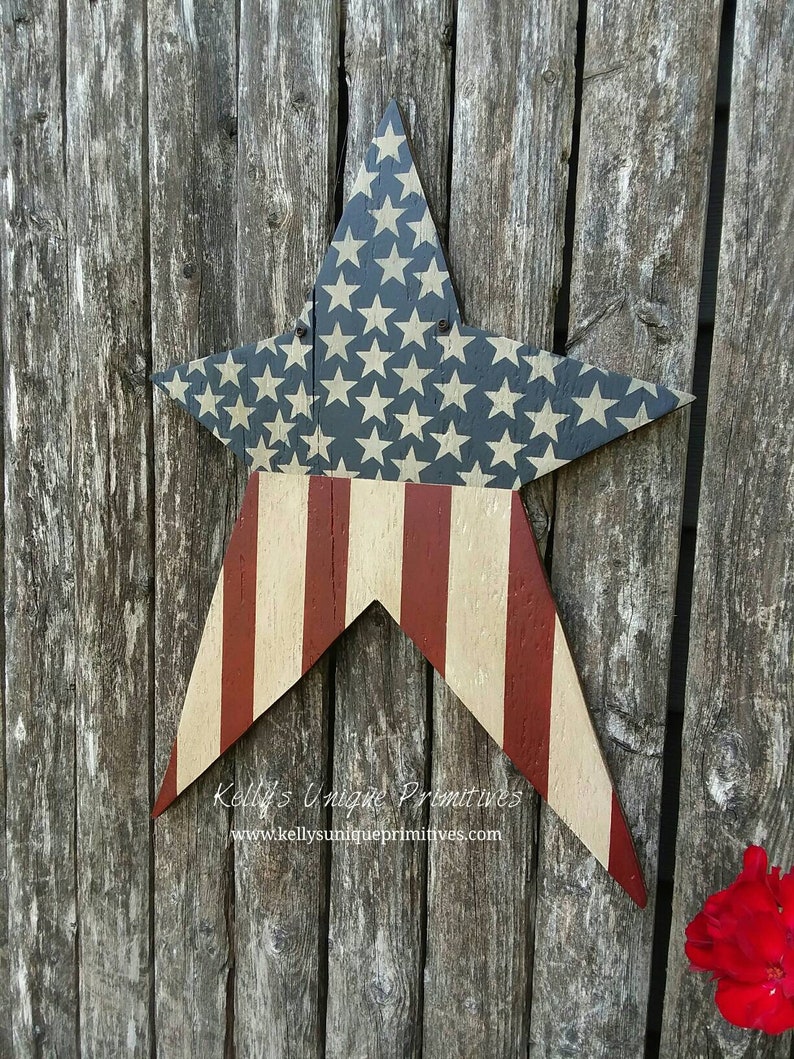 Op de afbeelding: Een houten stervormig bord geschilderd met de Amerikaanse vlag. De ster is geschilderd met rode en witte strepen en blauwe sterren op een witte achtergrond. Het bord hangt aan een houten hek. De tekst "Kelly's Unique Primitives" en "www.kellysunqiueprimitives.com" staat op het bord.