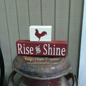 May include: Red wooden sign with white text that reads "Rise and Shine" with a white wooden block above it featuring a red rooster silhouette. The sign is sitting on top of a rusty metal milk can.