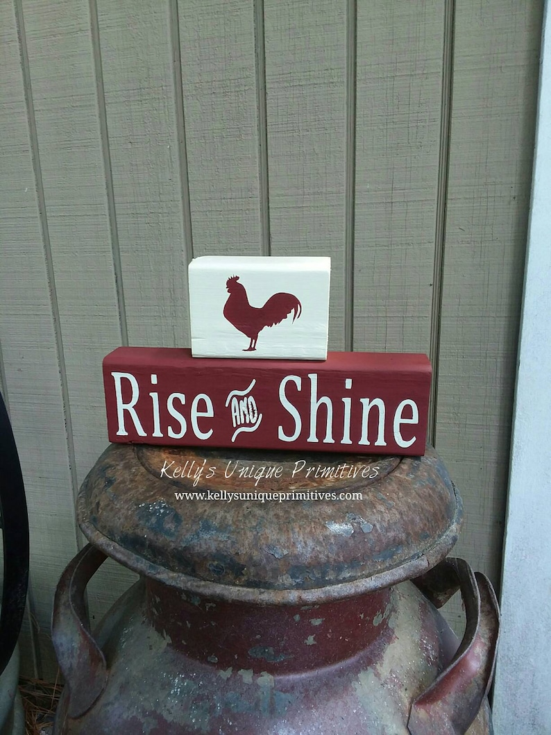 May include: Red wooden sign with white text that reads "Rise and Shine" with a white wooden block above it featuring a red rooster silhouette. The sign is sitting on top of a rusty metal milk can.