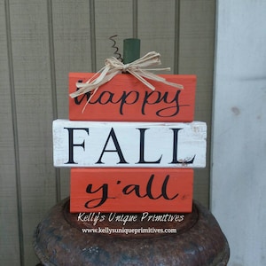 May include: Stacked wooden blocks painted in orange and white with the words "happy fall y'all" in black lettering. The blocks are arranged to resemble a pumpkin with a green and brown stem.