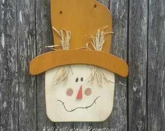 Peut inclure: Un visage de  l'épouvantail en bois avec un chapeau de paille, peint dans des tons de brun, blanc et orange. L'épouvantail a un visage souriant avec deux yeux noirs, un nez rouge et un sourire cousu.