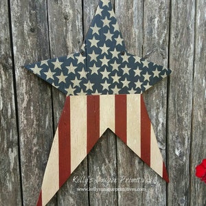 Op de afbeelding: Een houten stervormig bord geschilderd met de Amerikaanse vlag. De ster is geschilderd met rode en witte strepen en blauw met witte sterren. Het bord hangt aan een verweerd houten hek. De tekst "Kelly's Unique Primitives" en "www.kellysuniqueprimitives.com" staat op het bord.