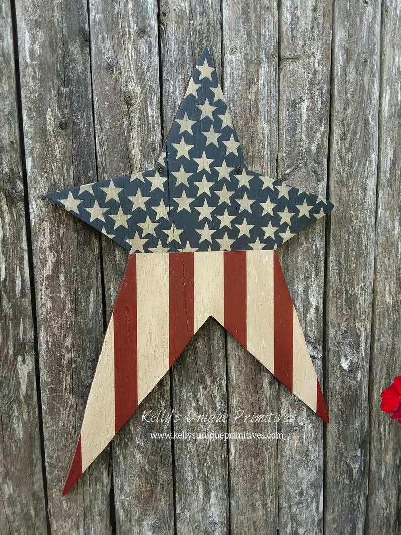 Op de afbeelding: Een houten stervormig bord geschilderd met de Amerikaanse vlag. De ster is geschilderd met rode en witte strepen en blauw met witte sterren. Het bord hangt aan een verweerd houten hek. De tekst "Kelly's Unique Primitives" en "www.kellysuniqueprimitives.com" staat op het bord.