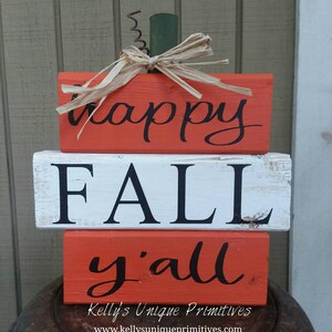 May include: Stacked wooden blocks painted in orange and white with the words "happy fall y'all" in black. The blocks are decorated with a straw bow and a green stem, resembling a pumpkin.
