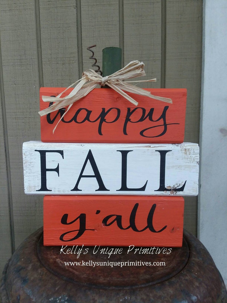 May include: Stacked wooden blocks painted in orange and white with the words "happy fall y'all" in black. The blocks are decorated with a straw bow and a green stem, resembling a pumpkin.