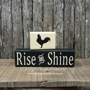 May include: Black and white wooden sign with the text "Rise and Shine" in white lettering. A smaller wooden block with a black silhouette of a rooster is stacked on top of the sign.