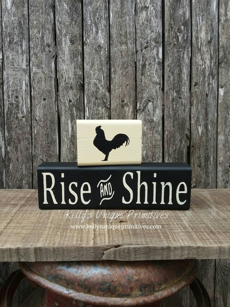 May include: Black and white wooden sign with the text "Rise and Shine" in white lettering. A smaller wooden block with a black silhouette of a rooster is stacked on top of the sign.