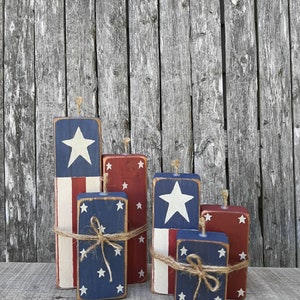 May include: Five wooden blocks painted in red, white, and blue with stars and stripes, resembling fireworks. The blocks are tied with twine and arranged in a staggered formation. "Kelly's Unique Primitives" and a website address are printed below the blocks.