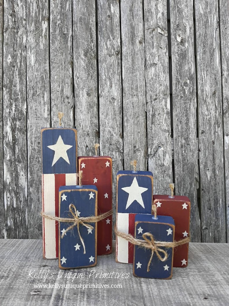 May include: Five wooden blocks painted in red, white, and blue with stars and stripes, resembling fireworks. The blocks are tied with twine and arranged in a staggered formation. "Kelly's Unique Primitives" and a website address are printed below the blocks.