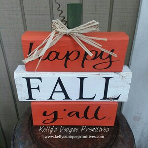 May include: A wooden pumpkin sign with the words "Happy Fall Y'all" painted in black. The pumpkin is stacked in three pieces and has a green stem and a straw bow.