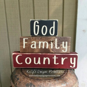 Puede incluir: Conjunto de tres letreros de madera apilados con las palabras "Dios", "Familia" y "Pa&iacute;s" pintadas en blanco sobre un fondo negro, marr&oacute;n y rojo. Los letreros est&aacute;n colocados sobre una superficie met&aacute;lica oxidada. El texto "Kelly's Unique Primitives" y una direcci&oacute;n de sitio web est&aacute;n impresos debajo de los letreros.