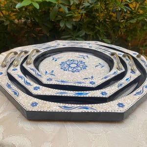 May include: Set of three decorative serving trays. Each tray is hexagonal with a black border, white mosaic-style surface, and blue floral accents. The trays have brass handles and nest inside each other.