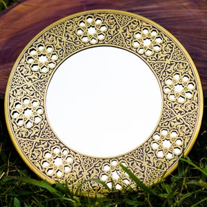 May include: A round, gold-colored mirror with an intricate black geometric pattern around the edge. The mirror is set against a backdrop of green grass and a dark brown wooden surface.