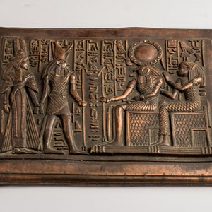 May include: A rectangular bronze relief depicting ancient Egyptian figures and hieroglyphs. The relief features detailed carvings of pharaohs, gods, and other figures, with a sun disc and other symbols. The artwork is likely a decorative piece.