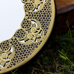 May include: A round mirror with a decorative gold and black border featuring an intricate geometric pattern. The mirror's edge has a dark brown finish. The background shows green grass and a wooden surface.