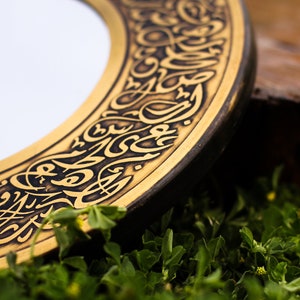 May include: Close-up of a decorative round object with gold and black accents. The object features intricate calligraphy around the edge. The background is blurred, with green foliage.