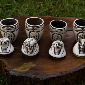 May include: A collection of white ceramic Egyptian-themed items. There are four cylindrical cups with black hieroglyphic designs and three small, decorative head-shaped pieces. The items are arranged on a wooden surface against a backdrop of green grass.