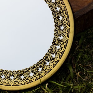 Può includere: Specchio rotondo con una cornice dorata ornata da un design traforato dettagliato. Il bordo dello specchio è marrone scuro e lo sfondo è un'area erbosa.