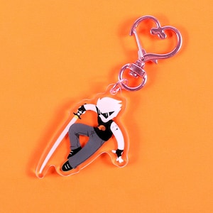 May include: A clear acrylic keychain with a cartoon figure in a black vest, grey trousers, and a sword. The keychain has a silver heart-shaped clasp and a silver ring. The background is orange. The figure has white hair.