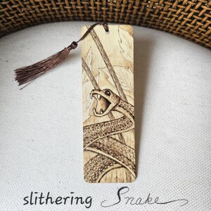 May include: Wooden bookmark with a detailed pyrography design of a snake with its mouth open. The bookmark has a brown tassel and the words "slithering Snake" are written below the image.