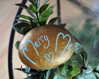 May include: A gold painted rock with the words "Marry Me" written in blue paint. The rock is surrounded by green plants.