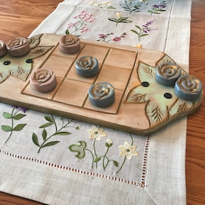 Może przedstawiać: Drewniana plansza do gry z siatką do gry w kółko i krzyżyk i rzeźbionymi detalami kwiatowymi. Plansza ma dziewięć pól z sześcioma ceramicznymi pionkami w kształcie róż. Trzy pionki są jasnoniebieskie, a trzy jasnoróżowe.
