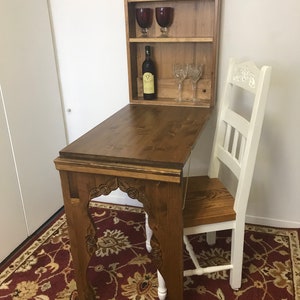 May include: A wooden wall-mounted wine bar with a fold-down table and a white chair. The bar has shelves holding wine glasses and a bottle of wine. The table and bar are brown, and the chair has a wooden seat and white painted frame.