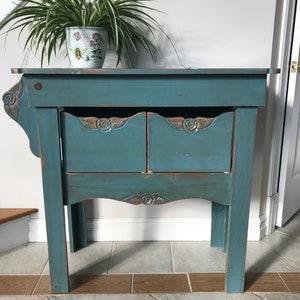 Peut inclure: Une table d'appoint en bois peinte en bleu sarcelle avec deux tiroirs et des détails floraux sculptés. La table a une finition vieillie et une plante en pot est posée dessus. La table mesure environ 76 cm de haut.