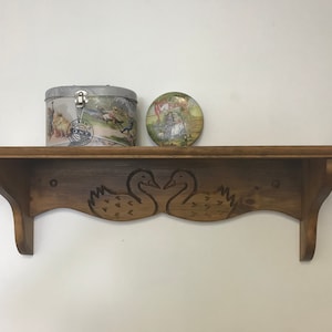 Puede incluir: Una estantería de madera con un diseño de cisnes tallados, que sostiene una lata vintage y un plato decorativo. La lata tiene una escena de carreras de caballos, y el plato presenta una imagen pastoral. La estantería está teñida de un color marrón cálido.