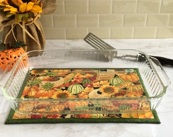 May include: A clear glass baking dish with a black handle sits on a green and yellow fabric hot pad with an autumn harvest theme. The hot pad features pumpkins, sunflowers, corn, and gourds.