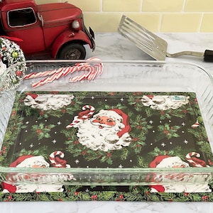 May include: A clear glass baking dish with a Santa Claus themed fabric hot pad underneath. The hot pad features a repeating pattern of Santa Claus faces, holly, and candy canes on a dark green background.