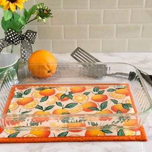 May include: A clear glass baking dish with a white and orange patterned fabric hot pad underneath. The hot pad has a design of orange slices and leaves.