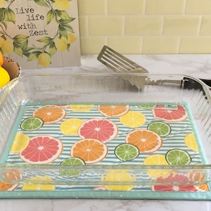 May include: A clear glass baking dish with a blue and white striped fabric liner featuring a citrus fruit pattern. The liner has a light blue border with white polka dots. The dish is sitting on a white countertop.