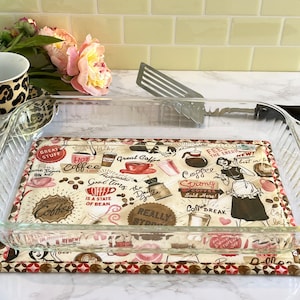 May include: A clear glass baking dish with a brown and white coffee-themed hot pad underneath. The hot pad features a variety of coffee-related illustrations and text, including "Great Coffee", "Coffee Strong", and "Coffee Break".