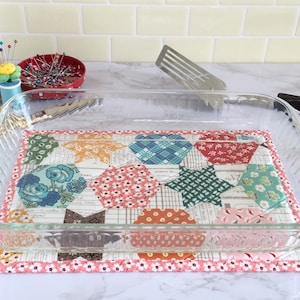 May include: A clear glass baking dish rests on a colorful quilted hot pad. The hot pad features a patchwork design with floral and geometric patterns in shades of blue, red, and orange. Sewing notions are in the background.