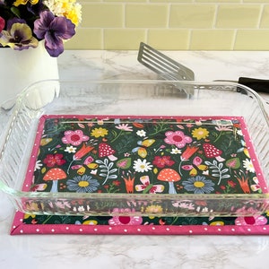 May include: A glass baking dish sits on a pink and white polka dot fabric hot pad with a floral print featuring mushrooms, butterflies, and flowers on a dark green background.