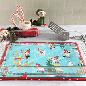 May include: A clear glass baking dish sits on a blue and red quilted hot pad with a Christmas gnome pattern. The hot pad has white polka dots on the red border.