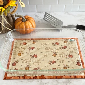 May include: A glass baking dish sits on a quilted kitchen hot pad with a fall-themed design featuring pumpkins and leaves in shades of orange, brown, and beige.