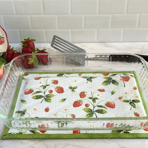 May include: A clear glass baking dish with a strawberry-themed quilted insert and a green polka-dot border. A ceramic strawberry-shaped container and a plate with strawberry designs are also visible, along with fresh strawberries and a spatula.