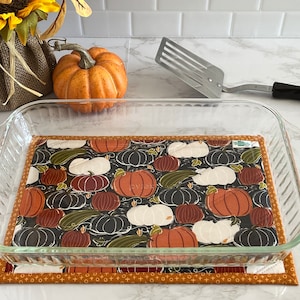 May include: A clear glass baking dish with a pumpkin-themed fabric hot pad underneath. The hot pad has a black background with orange, white, and green pumpkins.