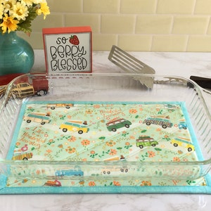 May include: A light blue quilted hot pad with a pattern of vintage vans and flowers. The hot pad is sitting on a white countertop with a glass baking dish on top of it. The text "Little adventures in a wonderful journey" is printed on the hot pad.