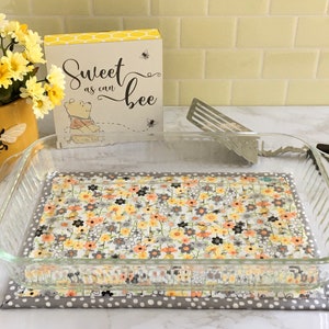 May include: A clear glass baking dish rests on a floral patterned trivet. The trivet has a gray polka dot border and a floral center with yellow, orange, and gray flowers. A decorative sign reads "Sweet as can bee."