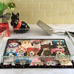 May include: A clear glass baking dish with a black and white fabric hot pad with a pattern of puppies wearing hats. The hot pad has a black border and colorful confetti.