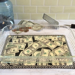 May include: A clear glass baking dish sits on a black and white patterned hot pad. The dish is filled with US dollar bills of various denominations.