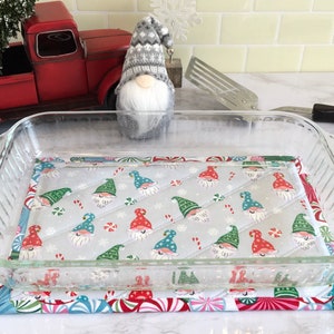 May include: A clear glass baking dish with a gray and white quilted hot pad with a gnome pattern. The hot pad has red, green, and blue accents.