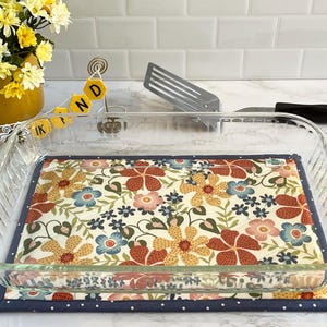 May include: A clear glass baking dish rests on a floral patterned pot holder. The pot holder features a cream background with orange, blue, and yellow flowers. A yellow banner with the letters "KIND" is visible.