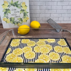May include: A glass baking dish with a lemon-themed fabric hot pad underneath. The hot pad has a navy blue background with white stripes and yellow lemon slices. The baking dish is on a wooden surface. The sign behind the dish says "Live life with zest".