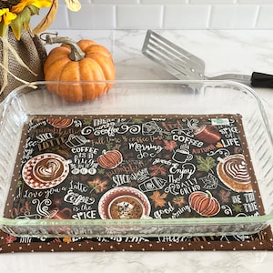 May include: A clear glass baking dish rests on a brown and black coffee-themed pot holder. The pot holder features coffee cups, pumpkins, and text like "Coffee is the Spice of Life." A metal spatula and a decorative pumpkin are in the background.